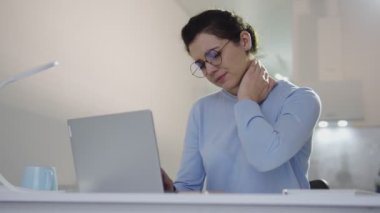 Kadın ofis çalışanı, dizüstü bilgisayarla çalışırken boynundaki ağrıyı hissediyor.