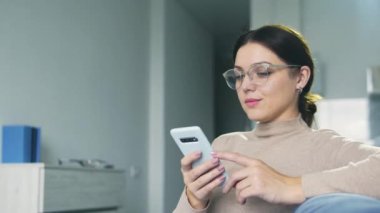 Çekici genç bir kadın akıllı telefonuyla kanepede oturuyor, arkadaşlarıyla sohbet ediyor ya da erkek arkadaşlarıyla mesajlaşıyor, yazışmalarını sürdürüyor ve gülümsüyor, aktif bir sosyal hayatı var.