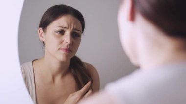 Üzgün genç kadın aynaya bakıyor, uzun kahverengi saçlarını tarıyor, parçalanmış ve kuru uçlarından memnun değil, bakım şampuanı ve saç kremini değiştirmesi gerekiyor.