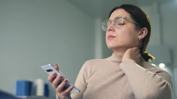 女性のオンラインマネージャは ソファの上に座って痛み首をマッサージし スマートフォンに入力します 鎮静ライフスタイルのために子宮頸神経のピンチや炎症 — ストック動画