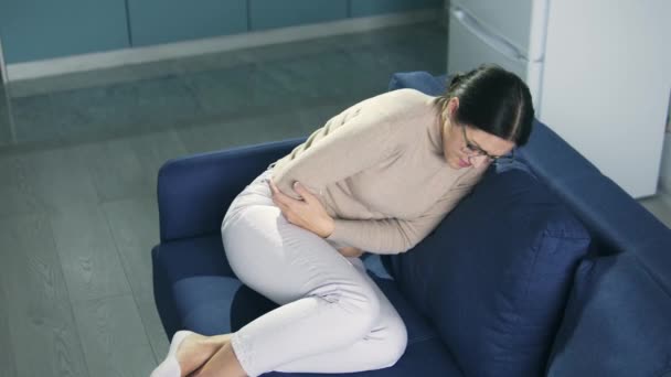 Молодая женщина лежит на диване, свернувшись в шар, чувствуя острую боль в желудке, предменструальный синдром, дисменорея, риск выкидыша или аборта, женское здоровье — стоковое видео