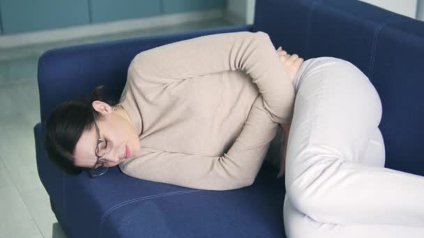 Deprimida mulher morena infeliz deitada no sofá e franzindo a testa de dor, de mãos dadas no estômago, sentindo dor aguda insuportável, risco de aborto, consequências do aborto — Vídeo de Stock