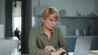 Kısa sarı saçlı ve şık gözlüklü bayan öğretmen öğrencilerin ödevlerini laptop 'da kontrol ediyor, sonuçlardan memnun değil, uzaktan öğrenme nedeniyle akademik performansı düşük.