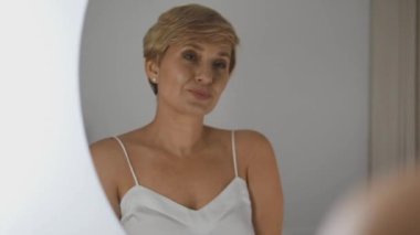 Yetişkin bir kadın aynaya bakarak eğleniyor ve yüzünü buruşturuyor.