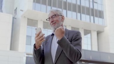 Mutlu, heyecanlı, gri saçlı adam dans ediyor. Akıllı telefondan iyi haberler okuyor, seviniyor.