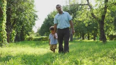 Çok ırklı baba ve tatlı kıvırcık saçlı oğul el ele tutuşup yüksek yeşil çimenlerde yürüyor, birlikte dışarıda vakit geçiriyorlar, babadan kalma izinler, bebeğe bakıyorlar.