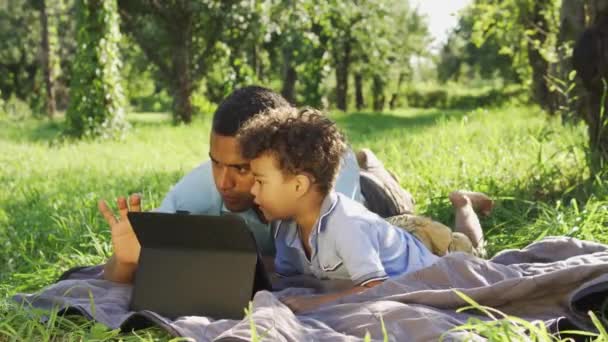パパはタブレットPcを使用して小さな出産の息子を教える 子供の運動能力と草の屋外に横たわっているメモリを開発するためにオンラインゲームをプレイする家族の男性メンバー — ストック動画