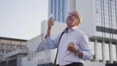 Başarılı, olgun bir ofis çalışanı evet işareti yapıyor ve dans ediyor, akıllı telefon ekranına bakıyor, bahis oynuyor, alışveriş yapıyor veya uygulamayı kolay teslimat için kullanıyor, faturaları internet üzerinden ödüyor, dijital ortama aktarıyor.