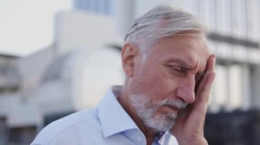 Beyaz gömlekli üst düzey bir ofis çalışanı güçlü bir rüzgarın üzerinde duruyor, yüzünü elleriyle gizliyor, gözlerini buruşturuyor ve kapatıyor, kafasında ağrı hissediyor, iç kulak iltihabı veya diş eti iltihabı riski, yüksek tansiyon