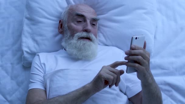 興奮した成熟した男性の入力メッセージスマートフォン ベッドに横たわって — ストック動画