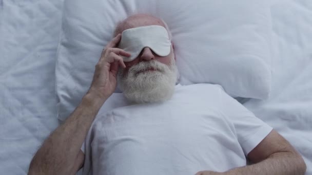 緊張した成熟した男性は ベッドに横たわって 睡眠の目のマスクを脱いで不快感を感じる — ストック動画