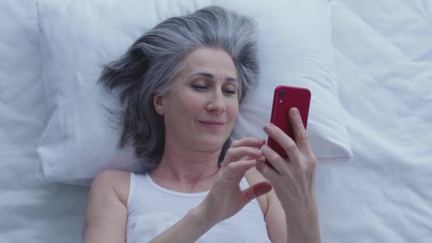 幸せな白髪の女性が使用してベッドでオンラインチャット日付アプリケーション インターネット — ストック動画