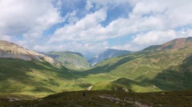 Yumuşak kıvrımlı bir dağ manzarasında yemyeşil bir vadi ve mavi gökyüzüne karşı bulutlar ve kameradan giderek daha az görünür hale gelen bir kız ön planda bir patikadan aşağı doğru yürüyor.