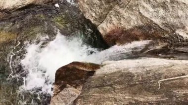 Yukarıdan gelen şelale, sağdan gelen su akıntısı büyük gri granit kayalar tarafından soldaki bir kaya havuzuna dökülüyor. Parlak hava kabarcıkları havuza giriyor.