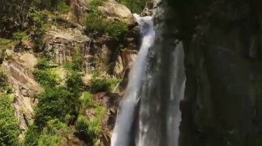 Gri-kahverengi granit kayalar ve yemyeşil orman arasında bir şelalenin yavaş çekim görüntüsü, kamera yukarıdan aşağıya suyla hareket ediyor turkuaz suyla dolu bir kaya havuzu ve spreyde süzülen bir gökkuşağı ortaya çıkıyor.