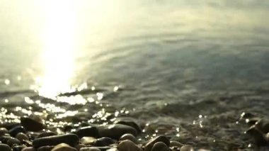 Ön planda çakıl taşları olan bir göl sahnesi, suyun üzerindeki güneşin yansıması odaklanmamış ve yumuşak altın bir ışık, ufka odaklanmadan ışığı takip eden kamera.