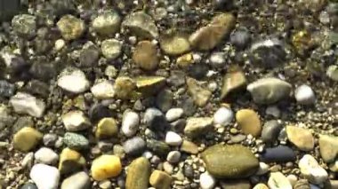 Yukardan çakıl taşlarıyla dolu bir göl kıyısı sahnesi. Küçük yumuşak dalgalar, odaklanmış çakıl taşlarının üzerinde düzenli olarak yıkanır.