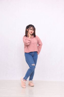 Gülümseyen, siyah saçlı, 20 'li yaşlarda, basit pembe tişörtlü, ayakta duran, boyun ve kalçalı güzel bir kadın servis promosyonu için beyaz arka plan stüdyo resmi.