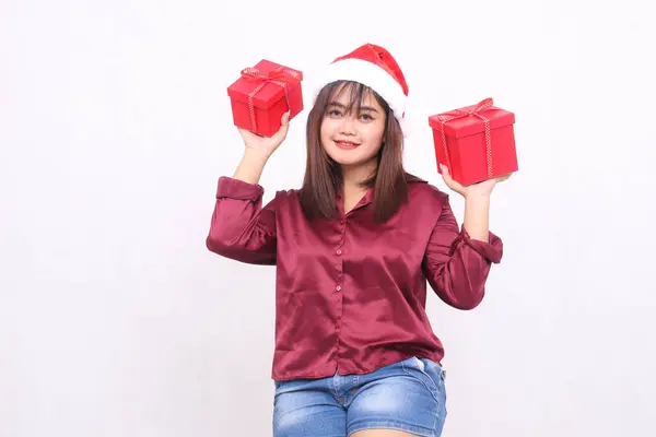 beautiful young asian indonesian girl smiling carrying gift box at christmas santa claus hat modern red shirt outfit lift box up on white background for promotion and advertising