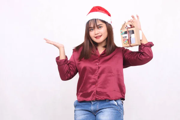 beautiful young woman southeast asian smiling left hand raised to gift hamper at christmas wearing santa claus hat modern red shirt outfit white background for promotion and advertising