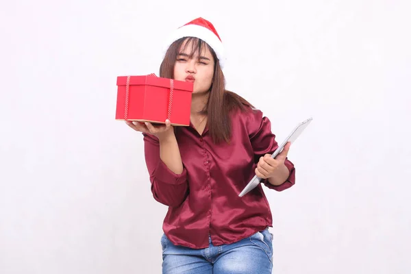 beautiful young southeast asian woman smile kissing hamper gift box and laptop tablet at christmas wear santa hat modern red shirt outfit white background for promotion and advertising