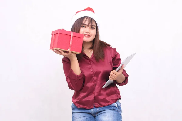 Beautiful young Southeast Asian woman smiling cheeks holding gift box hamper and laptop tablet at Christmas wearing Santa hat modern red shirt outfit white background for promotion and advertising