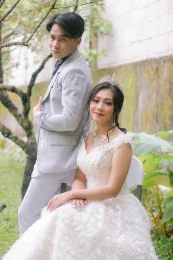 Bir çift zarif kadın ve erkek, rahat bir şekilde oturarak poz veriyorlar. Parktaki bir fotoğraf çekiminde güzel elbiseler giyiyorlar. Kameraya yakından bakıyorlar. Ulusal Gelin Açık havada düğün çifti