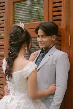 Zarif pozlar veren bir çift kadın ve erkek, resmi pozlar vererek tahta bir duvarın önünde duruyorlar. Parktaki bir fotoğrafta çapraz bacaklı güzel elbiseler giyiyorlar. Ulusal Gelin Açık havada düğün çifti