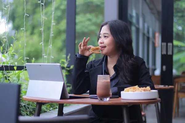 the elegance of a young Asian woman in a candid online meeting snacking in front of a laptop in a cafe