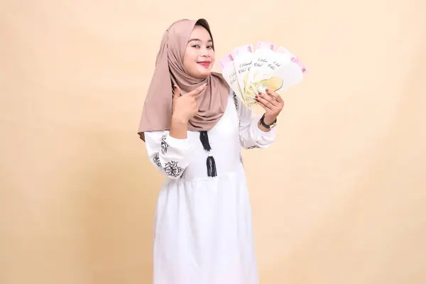 stock image portrait of a beautiful Asian Muslim woman wearing a hijab, smiling gracefully, holding and pointing to a gift or fitrah to the left on Eid day. used for advertising, giveaways, Eid and Ramadan