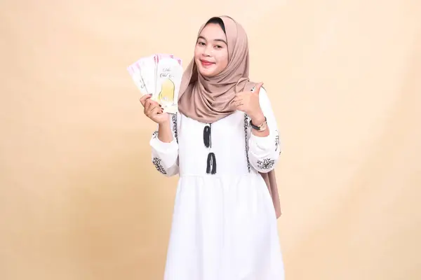 stock image portrait of a beautiful Asian Muslim woman wearing a hijab smiling with a thumbs up gesture holding a gift or fitrah to the right on Eid day. used for advertising, giveaways, Eid and Ramadan