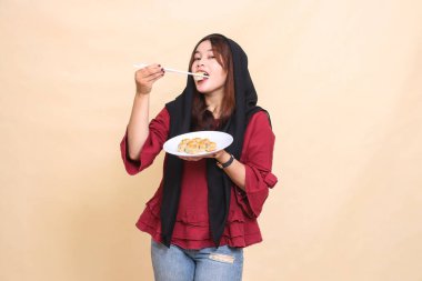 Kırmızı elbiseli güzel, olgun bir Asyalı kadın, başörtüsü takmış yemek çubuklarıyla gamsum yiyor ve içinde gamsum (Çin yemeği) bulunan bir tabağı kameraya bakıyor.