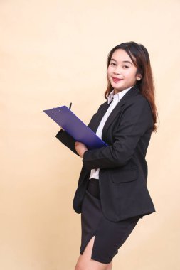 Güzel, resmi, Asyalı, yetişkin, takım elbiseli, gülümseyen ve notlar alan olgun bir kadın kameraya pano, ticari, boş ve iş yeri için sarı arka planda 3 'er 4 vücut portresi alıyor.