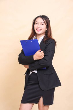 Takım elbiseli, güzel, resmi bir Asyalı yönetici kadın ayakta duruyor ve kameraya kalem panosu ile sarılıyor, ticari, boş ve iş yeri için sarı arka planda 3 'e 4 vücut portresi var.