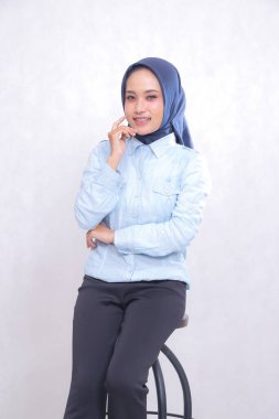 Genç Asyalı ofis kadını tesettürlü, mavi gömlekli, sandalyede oturuyor, neşeli bir şekilde gülümsüyor, kollarını kavuşturarak yanaklarını tutuyor. Güzel Müslüman kadınlar iş, finans ve moda içeriğini kullanır