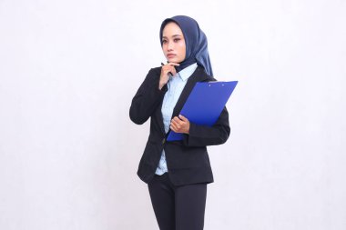 Endonezyalı tesettürlü bir ofis kadını elinde bir kalemle kameraya bakarak bir panoya sarılmayı düşünüyor. İş ve şirket için mavi gömlek giyen güzel Müslüman kadınlar.