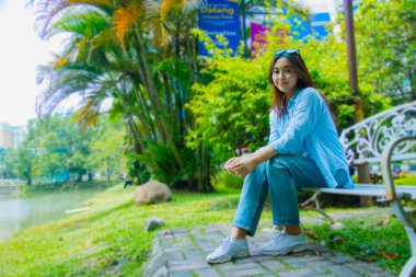 Güzel, gülümseyen Asyalı bir kadın kampüs parkında demir bir sandalyede rahatça oturuyor. İnsan ilgi alanı ve yaşam tarzı konsepti için öğrenci