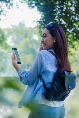 Güzel Asyalı kadın, güneşli yaz gününde kampüs parkında akıllı telefonlardan müzik dinlerken samimi bir gülümseme sergiliyor. Eğitim, tanıtım ve yaşam tarzı kavramları için kadın öğrenciler