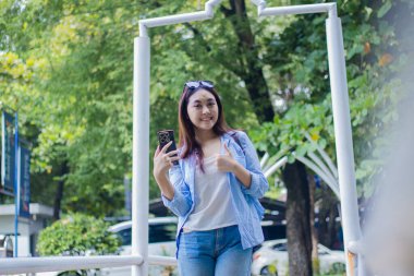 Neşeli, Endonezyalı, gözlüklü, akıllı telefon aleti takan güzel bir kadın güneşli havalarda kampüs parkında güzel bir jest yapıyor. Eğitim ve yaşam tarzı konsepti için bayan öğrenci