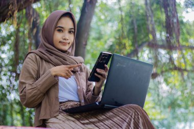 Endonezyalı güzel kadın zarif bir şekilde kameraya oturmuş parktaki dizüstü bilgisayarının önündeki akıllı telefon aletini işaret ediyor. eğitim, teknoloji ve yaşam tarzı konsepti için bayan öğrenci