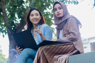 Endonezyalı bir çift güzel kadın, kampüs parkını işaret ederken dizüstü bilgisayar, kitap ve not panolarını taşıyarak samimi bir şekilde sohbet ediyorlardı. Yaşam tarzı kavramları için öğrenciler