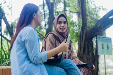 İki samimi Asyalı kadın kampüs parkında dizüstü bilgisayar, kitap ve dosya tutarak ders çalışırken gülümsüyor. Eğitim, tanıtım ve yaşam tarzı kavramları için kadın öğrenciler