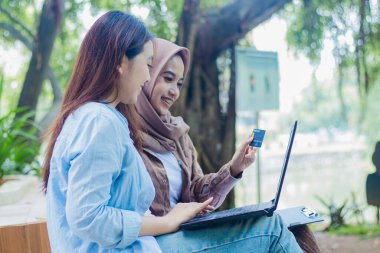 Bir çift mutlu Endonezyalı kadın, kampüs parkında ellerinde kalem, kitap ve pano tutarak online alışverişte indirim alıyorlar. Eğitim, tanıtım ve yaşam tarzı kavramları için öğrenciler