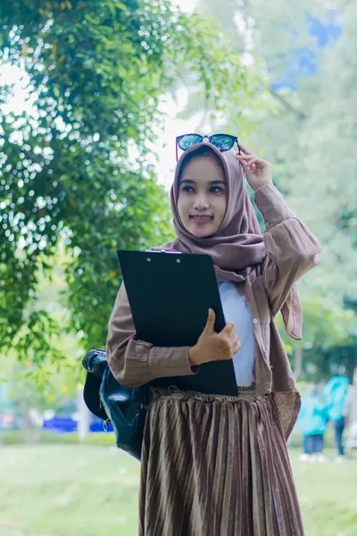mature asian woman in veil candid smile holding glasses carrying book clipboard shouldering bag standing in outdoor park. female students for the themes of education, technology and advertising