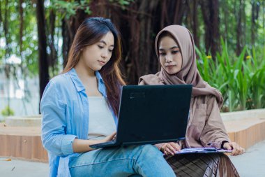 Bir çift Asyalı kadın kampüs parkında elinde kalem, kitap ve dosya tutarken dizüstü bilgisayara bakıyorlar. Eğitim, tanıtım ve yaşam tarzı kavramları için kadın öğrenciler