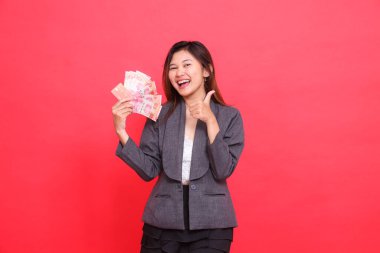 Elinde kredi kartı, banka kartı ve para olan mutlu bir Asyalı kadının, kırmızı arka planda gri ceket ve etek giyerek kameraya baş parmak işareti yapması. Reklam için.