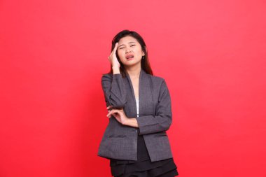 Kolları çaprazlanmış, başı dönmüş, başı dönen Asyalı bir ofis kadınının, elinde kamera, gri bir ceket ve kırmızı bir arka planı olan bir etek tutarken başı ağrıyor. Sağlık ve reklam için