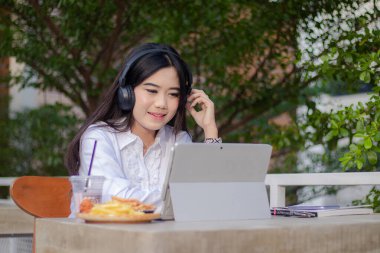 Ofis güzel Asyalı kadın açık hava kafede. Şımarık, gülen kız müzik dinlerken dizüstü bilgisayarın önünde çalışıyor içerik veya tanıtım için ön tarafa bakıyor.