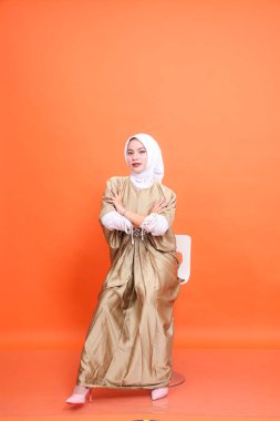 Kolları beyaz tesettürlü, kaftanlı, yüksek topuklu, turuncu arka planda izole bir sandalyede oturan Asyalı genç Müslüman kadının tam boy portresi. Çalışma anlayışı, moda