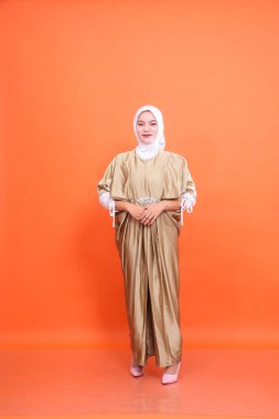 Tüm vücuduyla gülümseyen Asyalı Müslüman kadın resmi kaftan başörtüsü takıyor turuncu arka planda duruyor. İslam dini, moda ve ticari konsepti